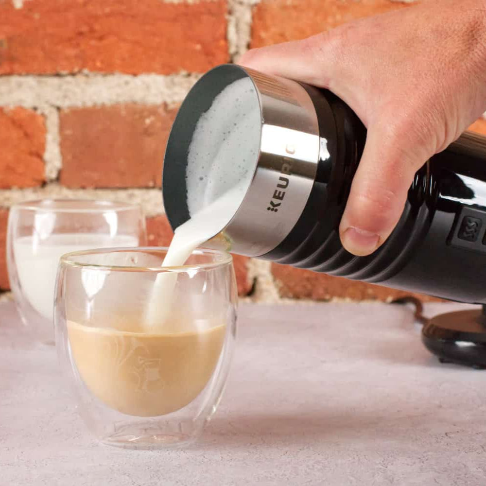 Hand pouring milk from a black and silver milk frother into a glass of coffee against a brick wall background.