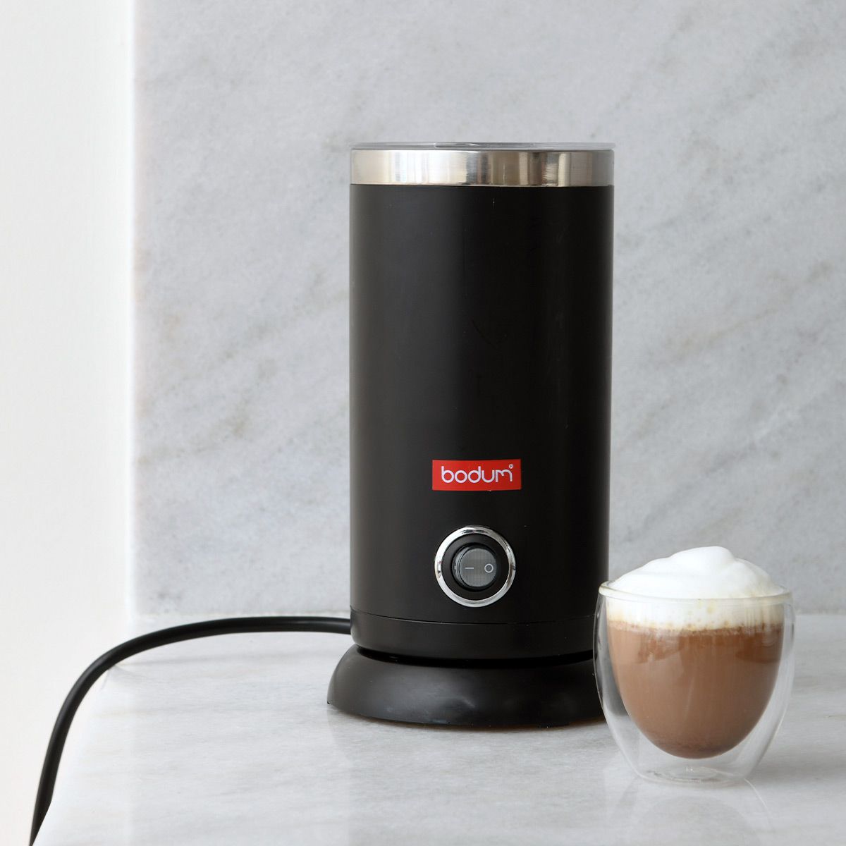 Black Bodum milk frother with a glass of frothed milk on a light gray background