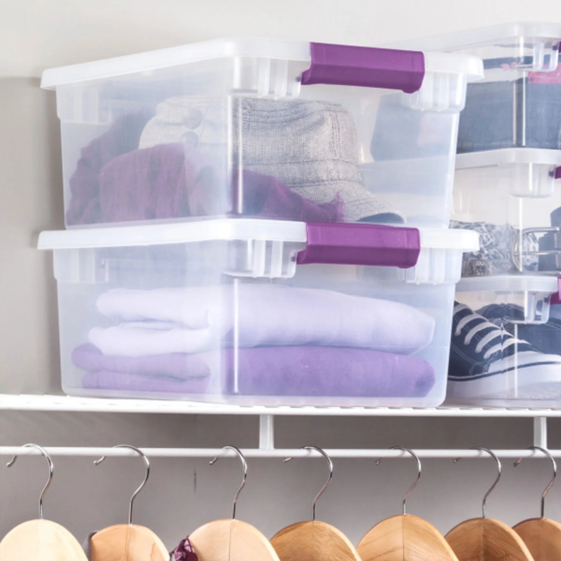 Clear storage bins with purple handles on a shelf above hangers