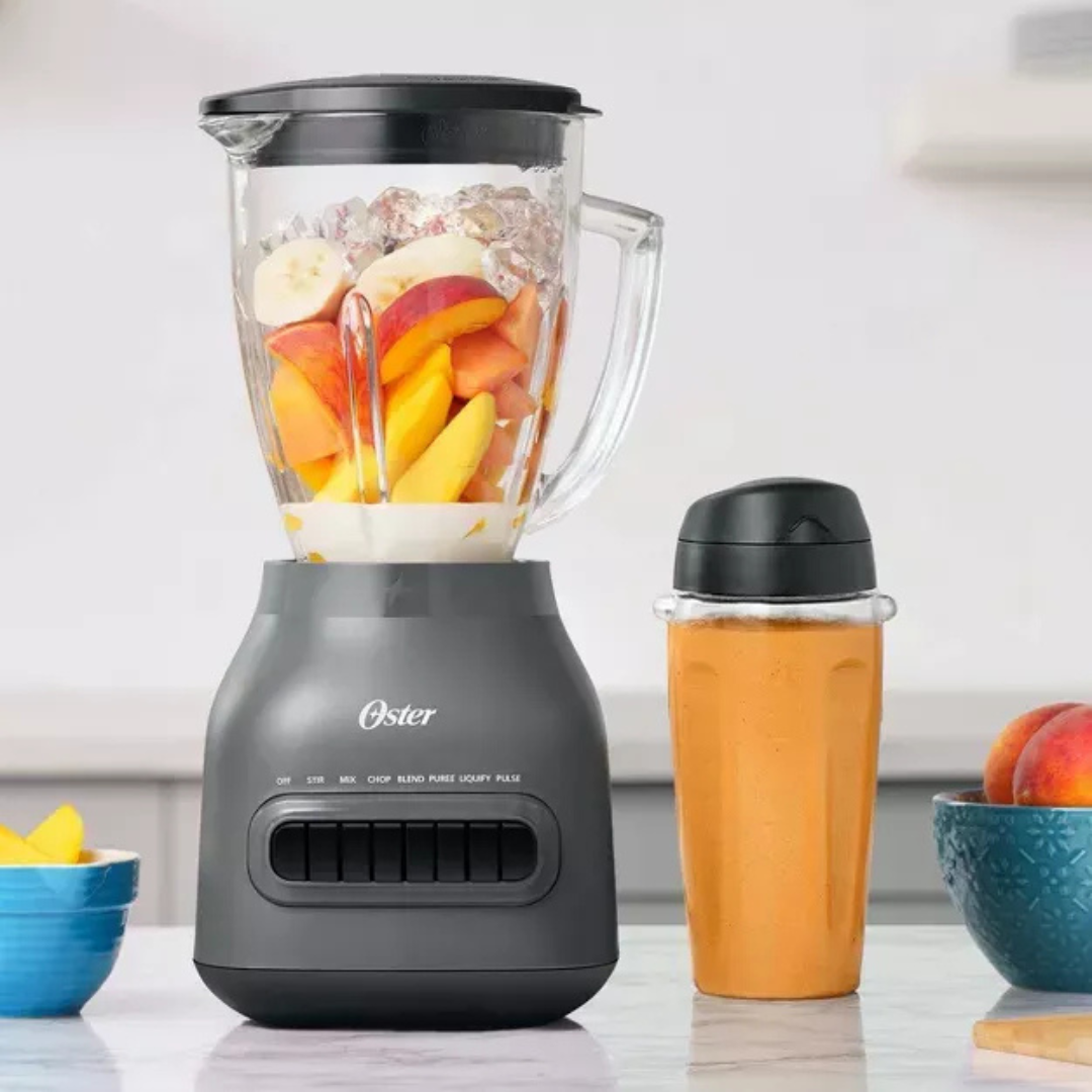 Oster blender with fruits inside on a kitchen counter, next to a smoothie and more fruits.