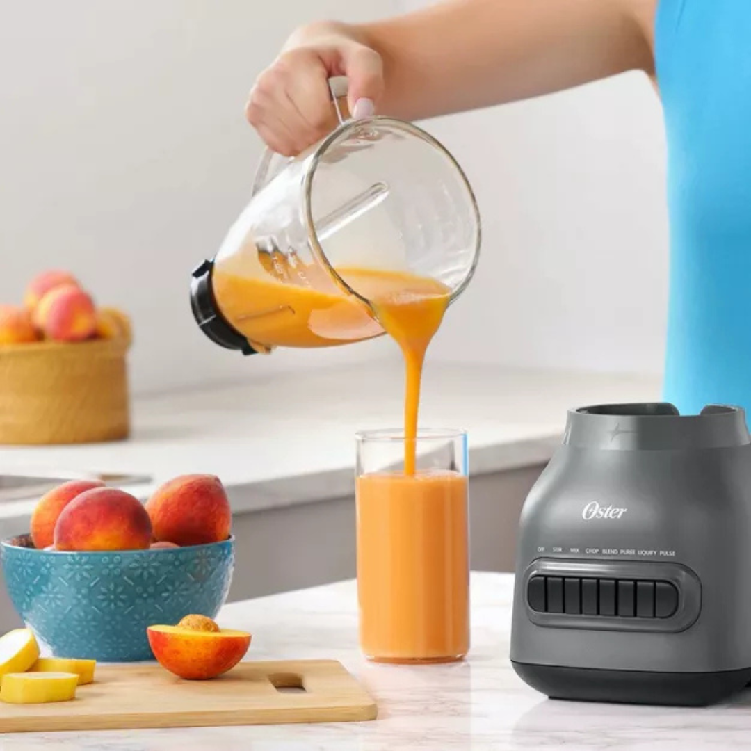 Person pouring orange juice from a blender into a glass, with a Oster blender base in the foreground.