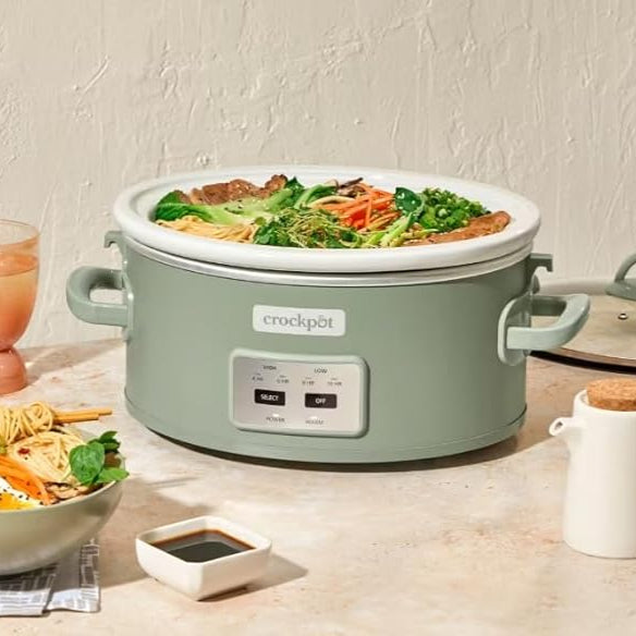 Crockpot with food on a table next to a bowl of salad and a glass of juice.