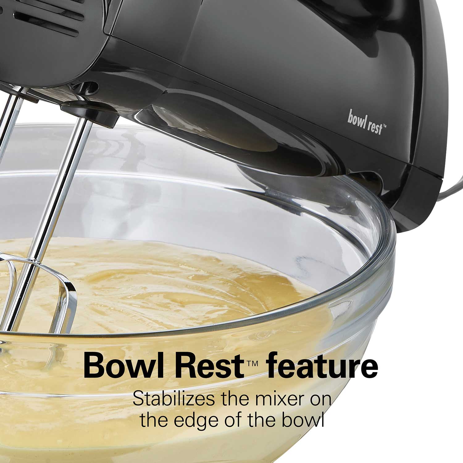 Black hand mixer with 'Bowl Rest' feature in action, mixing a yellow batter in a clear glass bowl.
