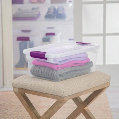 Stack of folded clothes in a clear storage box with a purple lid on a wooden stool.