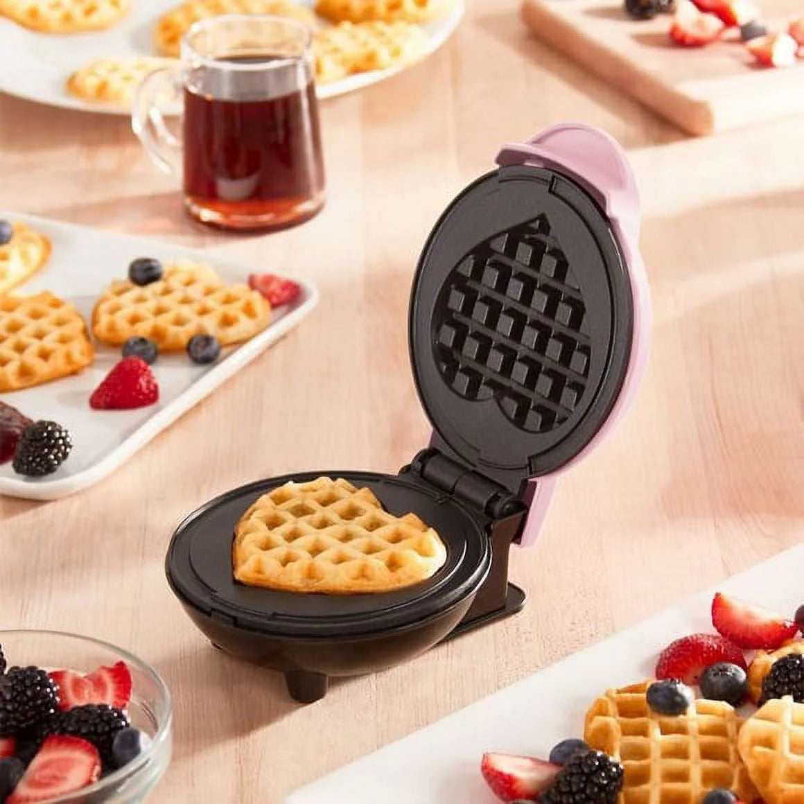 Heart-shaped waffle maker with a cooked waffle on a wooden table with waffles and berries.