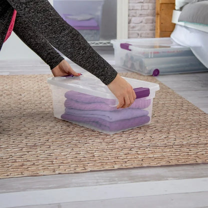 Person holding a clear storage container with purple towels on a wooden floor.