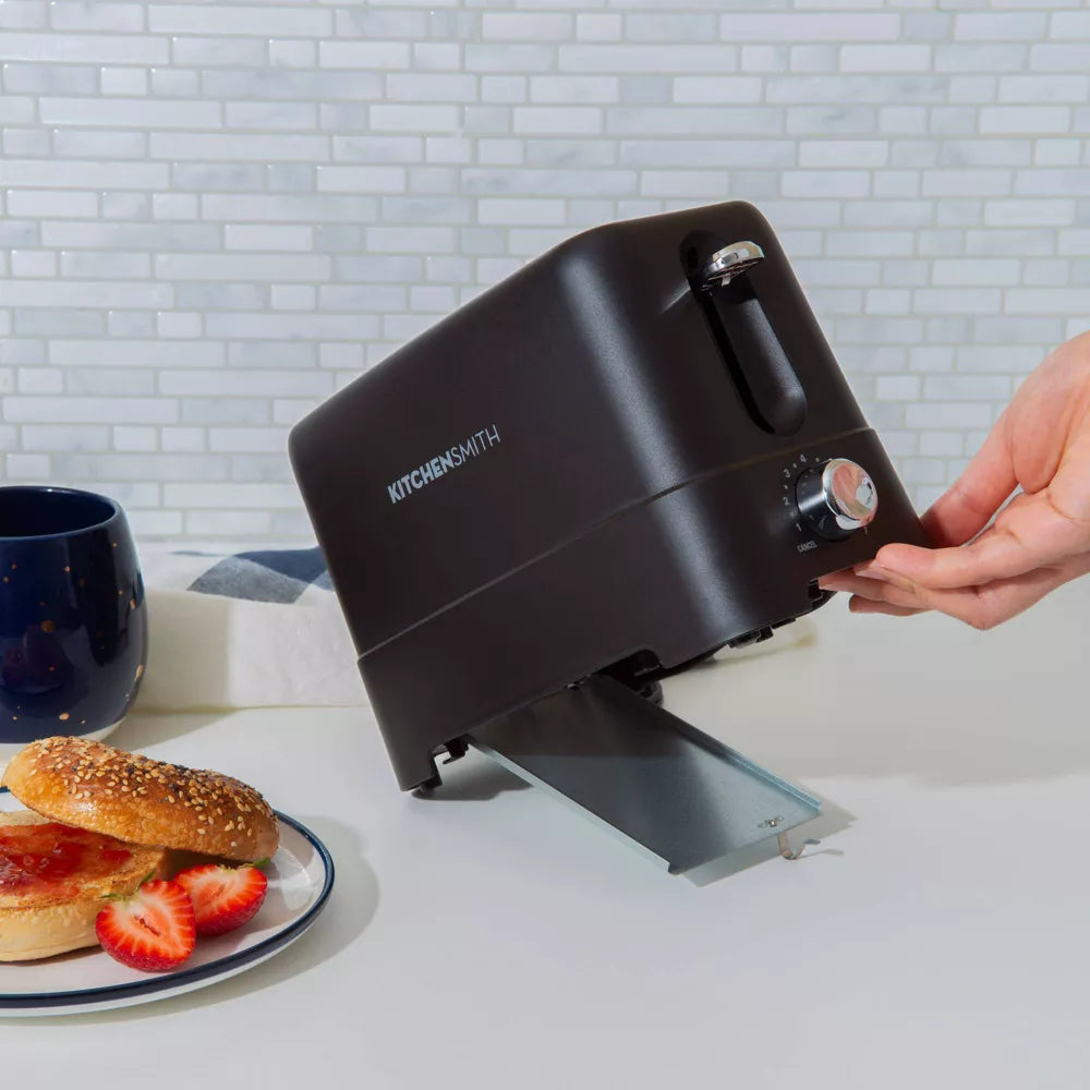 Black sandwich maker with a hand pressing down on it, next to a plate of sandwiches and strawberries on a kitchen counter.